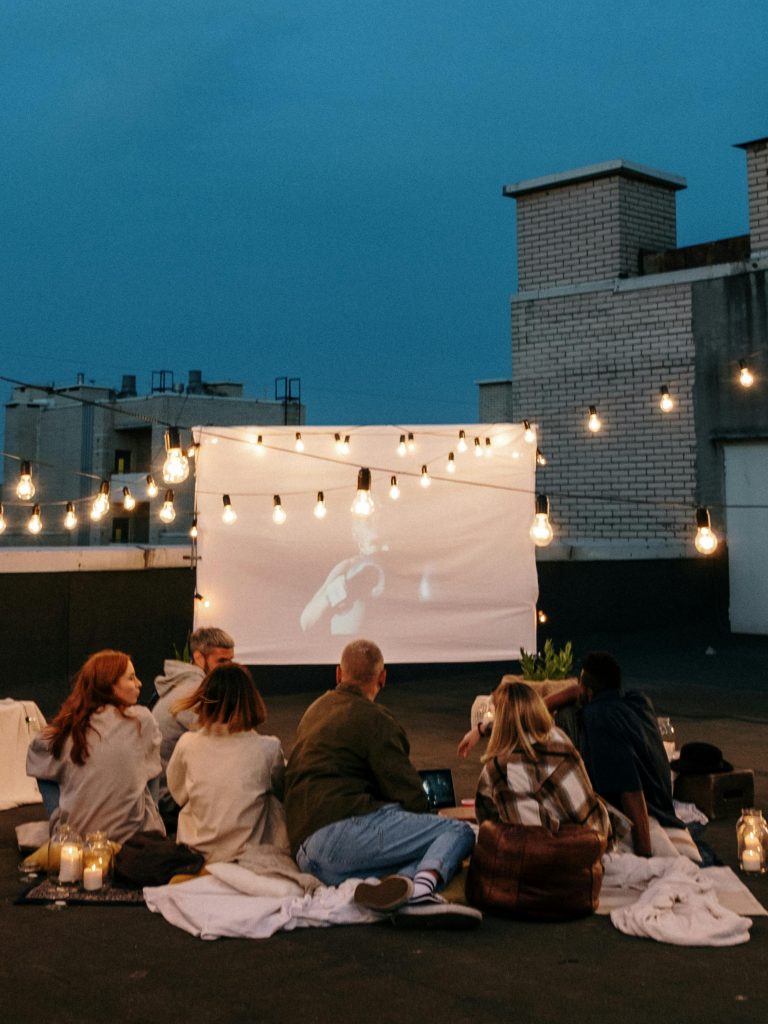 Menschen sitzen abends auf einer Dachterrasse vor einer Leinwand unter Lichterketten
